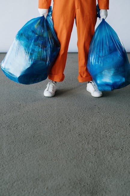 A person wearing a plaid shirt with shades of yellow, gray, and blue, along with a gray t-shirt underneath, is standing outdoors on a grassy area. They are holding open a large black rubbish bag lined with a silver inner layer, gripping the sides with both hands clad in bright green disposable gloves. The bag appears to be made of durable plastic with a slightly glossy finish, and it is partially open, ready to be filled with waste. The background consists of well-maintained green grass, suggesting the environment might be a garden, park, or outdoor space where waste collection or disposal activities are taking place. The scene aligns with a waste management or rubbish removal context, reflecting a straightforward approach to waste collection, possibly by an independent service such as Rubbish Collection Harringay, engaged in on-site clearance or private waste disposal. The lighting is natural, emphasizing the clarity of textures and colors in the scene, with no visible vehicle or additional equipment in view, focusing solely on the individual handling the rubbish bag.