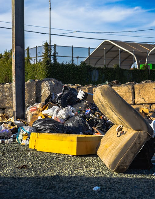 A narrow urban alleyway cluttered with various waste materials, featuring a large, heavy-duty black garbage bag in the foreground, positioned on the uneven asphalt ground that is stained and littered with small debris. Behind the bag, there are tall, graffiti-covered metal walls on the right and weathered brick walls on the left, with some graffiti tags visible. In the background, a pile of cardboard boxes, paper, and black plastic bags is stacked haphazardly against a wooden pallet, indicating a collection of refuse waiting for disposal or collection. Overhead, leafless tree branches extend across the scene, and a pale, overcast sky filters light, creating a muted, neutral tone. This setting illustrates a typical alleyway used for private waste handling, with discarded rubbish ready for collection, and Rubbish Collection Harringay's services implied in the clean-up context within this urban environment.