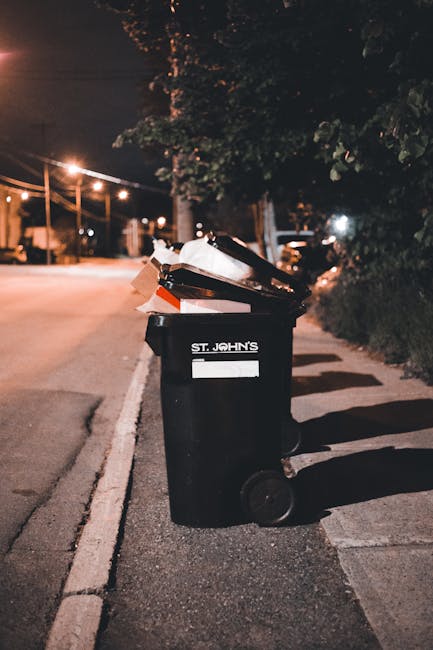 A black wheeled rubbish bin marked with 'ST. JOHN'S' in white lettering is positioned on a dimly lit urban sidewalk at night, with its lid open and filled with various waste materials, including a cardboard box, paper, and plastic items. The bin is situated near a curb, with shadows cast on the pavement from nearby streetlights and overhead trees with dark foliage. In the background, there are faint outlines of trees, streetlights, and parked vehicles, indicating a residential or mixed-use area. The scene captures a quiet moment of waste disposal or collection, aligning with the context of private rubbish handling and alternative waste management services such as those offered by Rubbish Collection Harringay. The lighting creates a subdued atmosphere, emphasizing the practicality and immediacy of rubbish disposal in an urban setting.