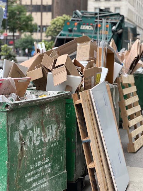 A collection of overflowing rubbish bins and waste scattered on a paved area in front of a commercial building with a blue scaffolding structure. The waste includes various black and red rubbish bags, flattened cardboard boxes, crumpled paper, plastic bottles, and discarded packaging materials, some spilling onto the ground. Behind the waste, there are two cars parked parallel to the pavement, partially blocked by a metal railing. The background shows storefronts with brightly colored signage and a pedestrian walkway, with two green bollards flanking the bins. The scene is illuminated by natural daylight, highlighting the cluttered and unkempt appearance of the waste, which underscores the importance of proper rubbish collection and disposal methods, such as those offered by independent waste removal services like Rubbish Collection Harringay.