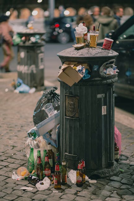 An overflowing public rubbish bin made of dark metal with vertical ridges and a rounded top, situated on a cobbled street surface. The bin's lid is open, revealing scattered paper and plastic packaging. Multiple discarded glass bottles with green and brown tones are placed around the base of the bin, some tipped over with litter on the ground, including crumpled paper and food wrappers. Resting on top of the lid are several takeaway drinks, including two cups with plastic lids and straws, and a paper coffee cup, along with a small container of food or condiment. The background shows blurred figures of pedestrians and parked vehicles, indicating an urban setting. A second rubbish bin is visible in the distance, along with some stray trash, suggesting littering and waste accumulation typical of street-side rubbish accumulation, highlighting issues related to inadequate waste management or informal disposal in a busy public area. The natural daylight emphasizes the gritty texture of the cobbles and the weathered appearance of the metal bin, aligning with private or independent waste collection scenarios often found in streetscapes.