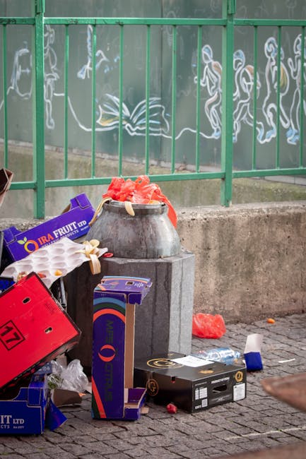 A metal rubbish bin with a rounded shape, placed on a concrete block, is overflowing with red and orange waste materials, possibly plastics or packaging debris. Around the bin are scattered cardboard boxes, one red with black and white markings, and another purple and blue box with a rainbow logo, indicating they contain waste or discarded products. The scene is set outdoors on a paved surface, with additional debris like a crumpled piece of paper or plastic, and a small piece of blue and white waste on the ground nearby. In the background, a green metal fence with vertical bars is visible, partially decorated with chalk or paint sketches of abstract figures and patterns on a mural or wall behind it, suggesting an urban environment. This scene exemplifies typical street-level rubbish accumulation that local waste collection services might handle or that private waste management companies such as Rubbish Collection Harringay could manage through alternative rubbish removal solutions.