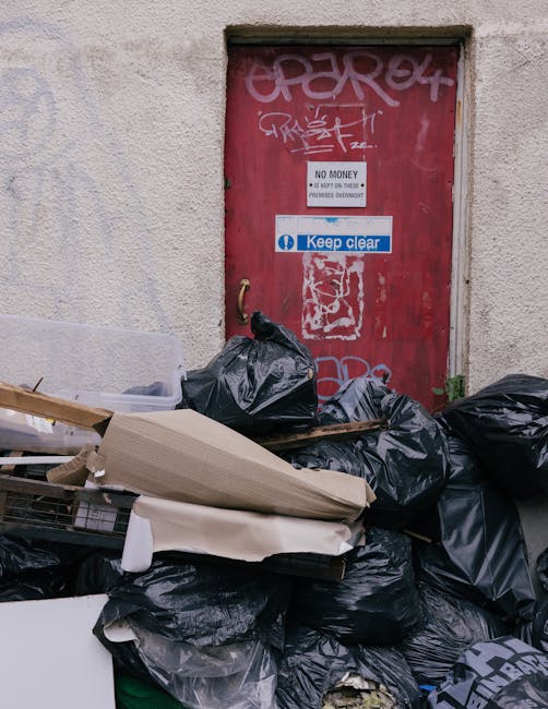 A collection of black rubbish bags and miscellaneous discarded items, including cardboard and packaging materials, piled up outside a red metal door set into a beige, textured wall. The door features graffiti tags, a white sign reading 'NO MONEY,' and a blue and white 'Keep clear' sign. The bags and debris occupy the foreground, partially obstructing the lower portion of the door. The scene appears to be an outdoor urban area, possibly a back alley or service entrance, with the waste awaiting collection or removal. The overall environment suggests a situation where independent rubbish clearance, such as that offered by Rubbish Collection Harringay, might be required to manage waste in this space.