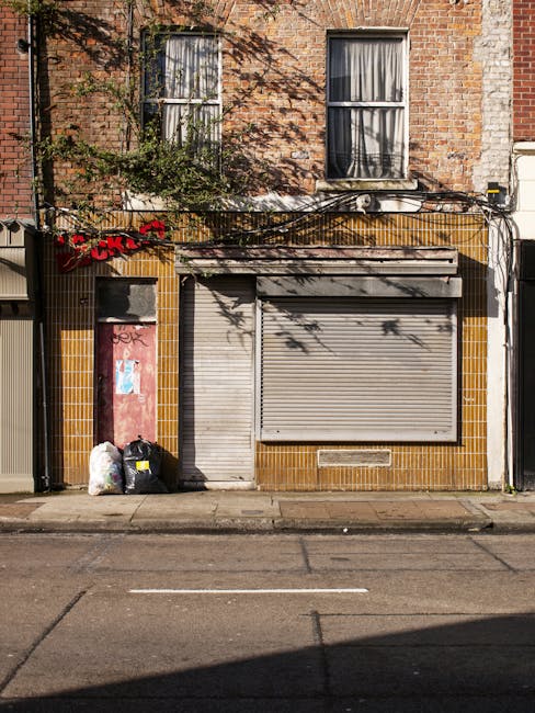 The image depicts the exterior of a brick building with an aged, textured facade, featuring two large, vertically elongated windows with white frames and partially open curtains in the upper section. The ground level shows a closed metal roller shutter painted in a light grey tone, flanked by a small, worn door on the left side, partially covered with graffiti and weathered paint. Adjacent to the door, there are two black trash bags and a white plastic bag, placed on the pavement in front of the building, suggesting an area of rubbish collection or disposal. Above the ground floor, black plastic cables and wiring run horizontally across the facade between the windows, casting shadows on the brickwork. The building’s exterior surfaces include sections of yellowish-brown tiles around the lower storefront area, contrasting with the natural brickwork. The scene is illuminated by daylight, with some natural shadows from nearby objects, creating a realistic view of an urban waste management scene involving private rubbish collection, as managed by Rubbish Collection Harringay, in an area where the building’s face shows signs of age and recent waste disposal activity.