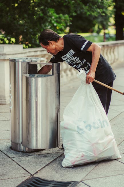 A middle-aged man with dark hair, wearing a black T-shirt with white and blue text and black trousers, is seen disposing of waste into a stainless steel public litter bin on a paved sidewalk. He is holding a large white plastic bag filled with rubbish, which rests on the ground beside him, and is leaning forward to deposit waste into the bin. The scene is set outdoors in a green area with trees and bushes in the background, and a low stone or concrete fence running along the edge of the pavement. The image captures a moment of urban litter collection or waste disposal, with the man engaging in personal waste management as part of maintaining a clean public space, which reflects themes of private rubbish disposal and alternative waste handling methods often supported by local waste management services like Rubbish Collection Harringay in the N4 area.