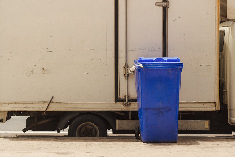 A blue wheeled rubbish bin with a closed lid is positioned on a pavement next to a white delivery or storage truck with a plain, slightly worn surface. The bin has a glossy plastic finish with some dirt and scuff marks visible on its surface. It is placed directly in front of the truck’s side panel, which appears to be made of metal and shows faint scratches and dirt marks. The truck's rear wheel is partially visible beneath the elevated side panel, indicating it is parked on an urban sidewalk or driveway. The environment around the scene is lit with natural daylight, suggesting daytime conditions, and the background is out of focus, emphasizing the bin and vehicle as the main objects. This setting reflects a typical scene involving rubbish collection or disposal, commonly associated with independent waste handling services such as those offered by Rubbish Collection Harringay for managing bulky waste or on-site clearance, especially in areas like Harringay Ladder.