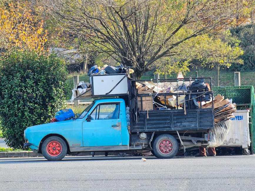 A small, old blue pickup truck is parked on the side of a street, heavily loaded with various types of rubbish and waste materials. The truck's body is made of metal with visible signs of wear, rust, and chipped paint, especially on the door and around the wheels. The cargo area, which is metal-framed and open, is filled with a mixture of cardboard boxes, plastic bags, and other debris, some tied with ropes or straps to secure the load. A large white appliance or piece of furniture is situated towards the front of the cargo bed, partially covered by black plastic bags. The van's red steel wheels contrast with the blue paint, and the vehicle is positioned next to a curb with a well-maintained hedge on one side and a green metal fence behind, with a large tree whose branches and leaves extend overhead, creating a dappled sunlight pattern. The environment suggests an area where private rubbish collection or on-site clearance, possibly by a waste management service such as Rubbish Collection Harringay, might be involved, supporting alternative waste handling methods outside of municipal collection.