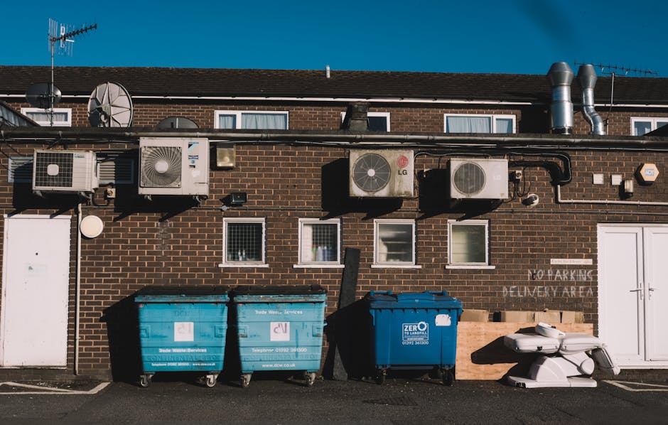 The image shows the rear exterior of a brick building under a clear blue sky, featuring a row of five wall-mounted air conditioning units of varying sizes and brands, positioned along the upper section of the wall. Below the units, three large waste bins—two blue and one dark blue—are situated in the foreground, all with visible wheels and lid closures, and branded with waste management company logos. Adjacent to the bins, there are some white plastic debris or furniture parts stacked on the pavement, indicating on-site rubbish clearance. The building has three small rectangular windows with white frames, with some having blinds or curtains behind them, and a white double door on the right side. To the right of the bins, there's a faded 'NO PARKING DELIVERY AREA' sign painted on the wall, reflecting restricted access in that area. The setting appears to be a typical urban or suburban back alley used for waste disposal and external service access, with natural daylight illuminating the scene, consistent with private rubbish collection or alternative waste handling arrangements managed by local waste services such as Rubbish Collection Harringay.