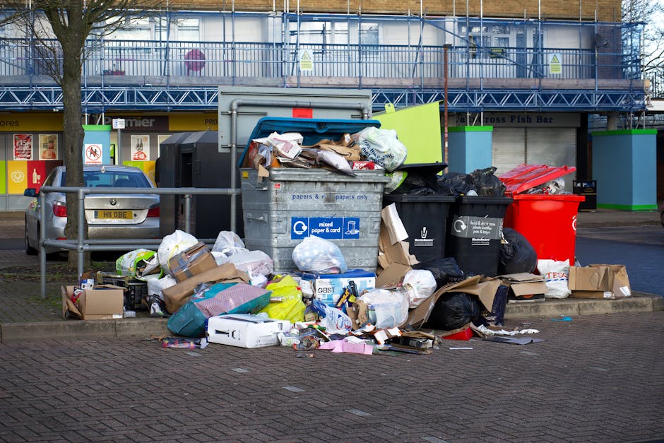 Image showing a large accumulation of mixed waste debris situated on a paved section of an urban street, with various black, black, and red rubbish bags, along with open cardboard boxes and loose packaging materials, spilling over a metal barrier into the surrounding area. The waste comprises paper, cardboard, plastic bags, and packaging materials of different colors and textures, some flattened and others crumpled, indicating previous disposal activities. Behind the rubbish pile, a grey commercial waste bin with a blue label designated for mixed paper and card is visible, partially filled with untidy waste overflow. To the left, a parked silver car with a yellow license plate is positioned adjacent to the waste area, and in the background, there are storefronts and a building under construction with blue scaffolding and safety signage. The scene is illuminated by diffuse daylight, highlighting the disorganized state of the refuse, which appears to be awaiting collection or disposal, characteristic of private waste management services such as those provided by Rubbish Collection Harringay, serving the local area with alternative waste handling solutions.