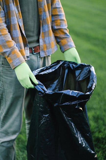 A person wearing a checked shirt with shades of yellow, blue, and gray, over a dark green T-shirt, is standing outdoors on a grassy area. They are holding open a large black rubbish bag with both hands, which has a glossy finish and appears to be made of heavy-duty plastic. The individual is also wearing light green gloves, indicating they are engaged in waste collection or disposal activities. The background shows a well-maintained lawn with short, green grass, suggesting a park or garden setting. The scene reflects an activity related to rubbish removal or cleaning, with a focus on responsible waste handling. The lighting suggests natural daylight, highlighting the textures and colours of the clothing and rubbish bag. This image aligns with the context of independent waste collection services such as those provided by Rubbish Collection Harringay, supporting on-site rubbish clearance in a professional manner.