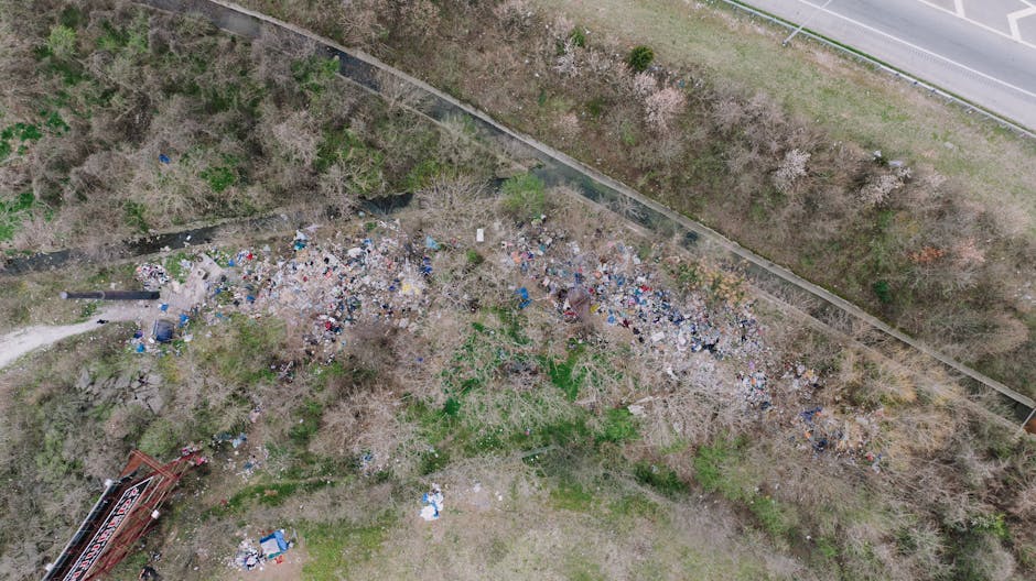 An aerial view shows a large, irregularly shaped collection of accumulated litter and debris spread across a patch of land near a fence and a pathway, with various discarded items including plastic bottles, bags, and paper scattered among leafless trees and sparse vegetation. To the right, there is a paved road with a grassy verge separating it from the affected area. The waste appears untreated and unmanaged, indicating a need for proper rubbish removal services such as those provided by Rubbish Collection Harringay, which helps manage waste in residential or neglected outdoor areas. The scene illustrates a typical site where private waste collection or on-site clearance might be necessary, highlighting the importance of adhering to local council waste disposal regulations while considering alternative rubbish handling methods. Natural light illuminates the scene, revealing the textures of litter contrasting with the surrounding greenery and bare branches, emphasizing the unmanaged state of this environmental space.